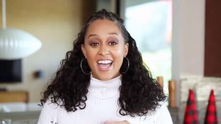 Tia Mowry in a white shirt with curly hair smiling into a camera.