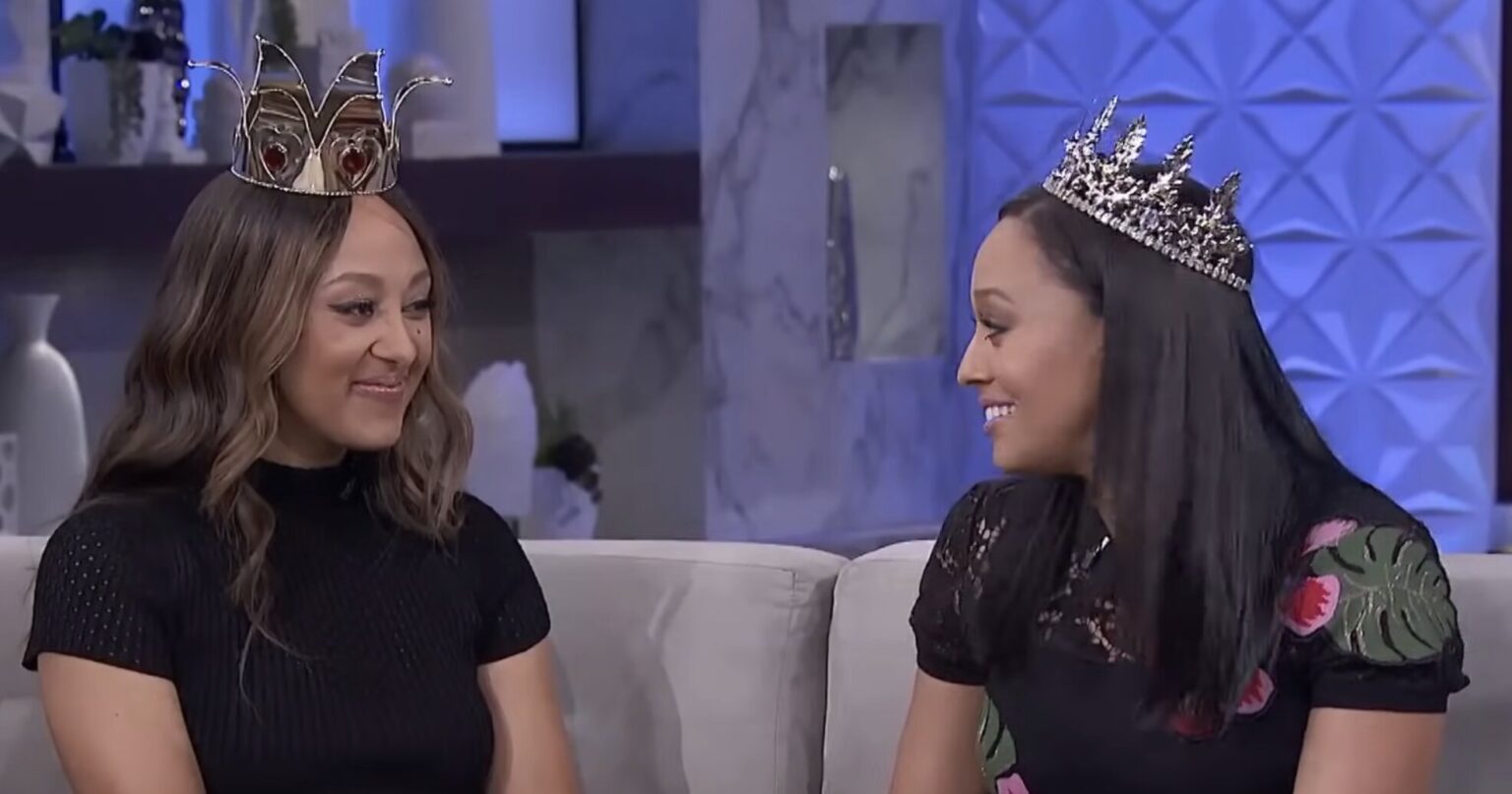 Tia and Tamera Mowry sitting on a couch with crowns on their heads.