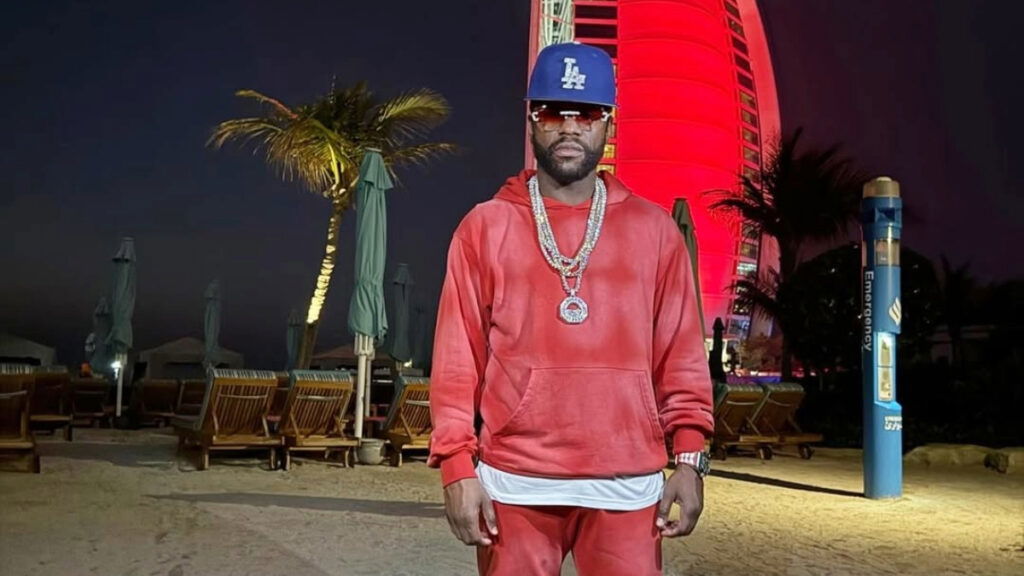 Floyd Mayweather in a red sweatshirt, pant and blue facecap standing in a dark street.