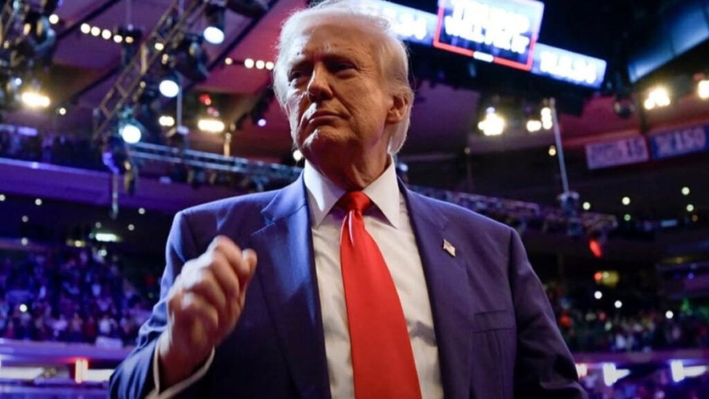 A close-up of Donald Trump with a crowd behind him and his name on a giant screen above