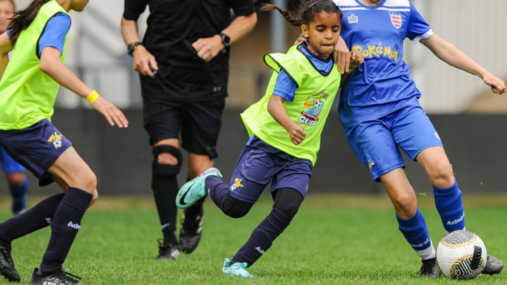 Kids wearing uniforms with Pokemon logos play soccer, one child dribbling while another swoops in for a steal