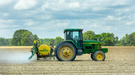 A green tractor drives across a field of dirt, leaving a trail behind it